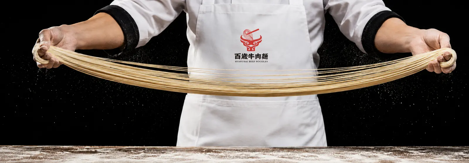 百歳牛肉麺の牛頭と麺鉢のロゴ入り白いエプロンを着けた職人が、両手で長い手延べ麺を伸ばす様子